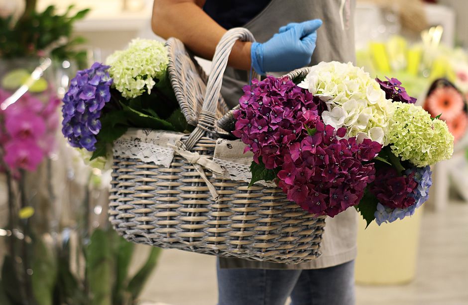 A young delivery person wearing a red cap and a light-colored uniform holds out a bouquet of white flowers towards the camera, smiling warmly. The bouquet is composed of fresh, vibrant white blooms wi