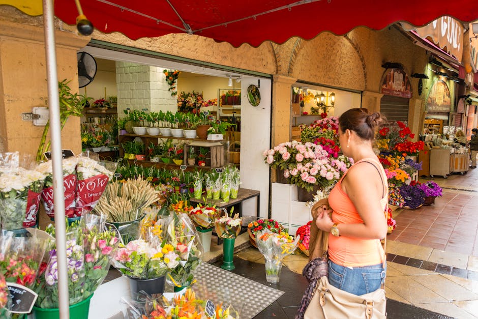 A woman standing in front of an outdoor flower shop on Bromley High Street, browsing a vibrant display of fresh flowers including pink roses, purple chrysanthemums, red carnations, and mixed floral bo
