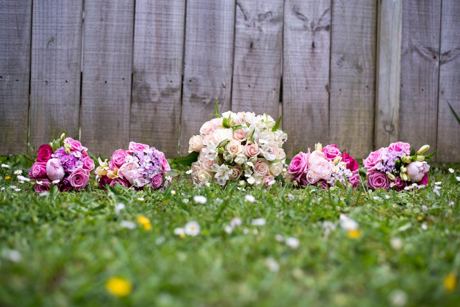 Multiple floral arrangements composed of pink, white, and cream roses, and purple and white hydrangeas, arranged in small bouquets on a grassy surface in front of a weathered wooden fence. The bouquet