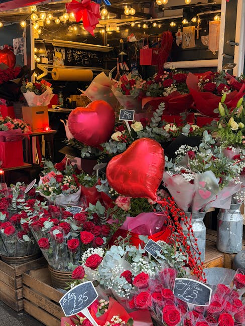 A display of various floral arrangements featuring red roses, white baby’s breath, and greenery, arranged in baskets and wrapped in pink and white paper, with prominent red heart-shaped balloons amo